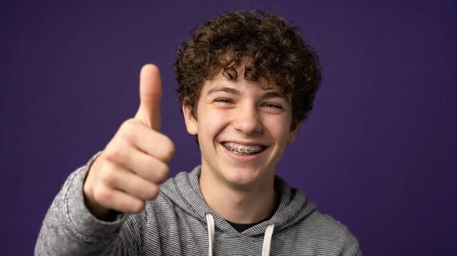 A teenager with braces smiling and showing thumbs up on a purple background
