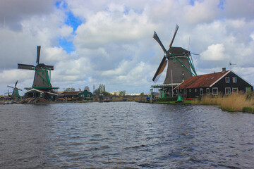 Naklejka premium May 20 2025 Zaanse Schans Netherlands Traditional Historic Wooden Windmills on the Riverbank Under Cloudy Blue Sky