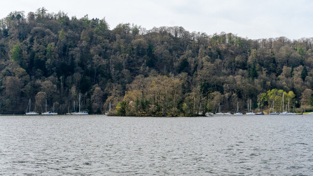 Lake Windermere in the Lake District National Park, where one of the picturesque, small, wooded islands sits in the center of the frame, surrounded by boats and sailboats near the forested shores