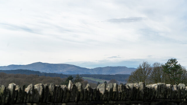 A traditional dry stone wall in the foreground with rolling hills, woodland, and distant fells or mountain ridges under a partly cloudy sky
