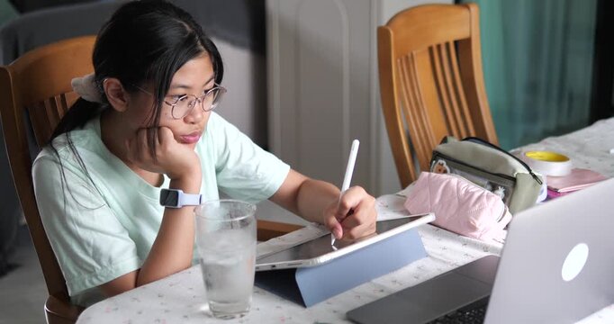 Asian teen girl resting chin on hand studying online with digital tablet at home