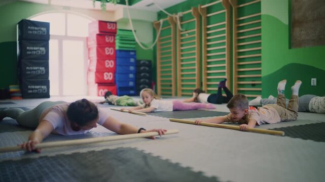 A group of children and adults performs fitness exercises with wooden sticks on the gym floor. Young people and an instructor engage in physical training, building core strength.