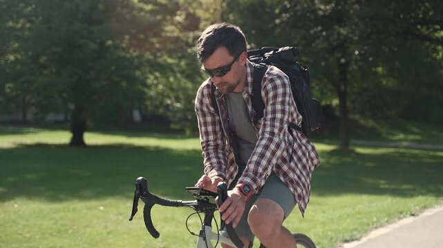 Distracted man in a plaid shirt riding a bicycle in a green city park while looking down and using his smartphone as a GPS navigator on the handlebar creating a very dangerous traffic road hazard