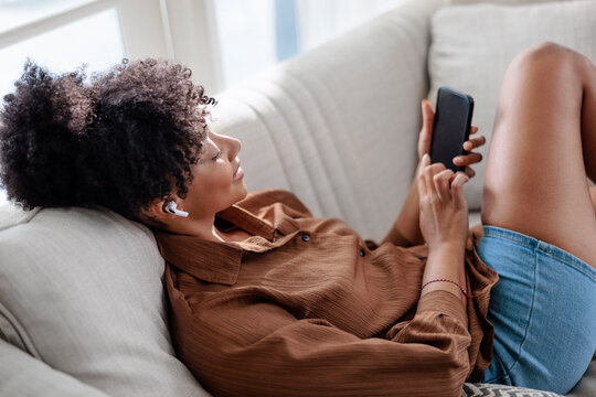 Woman relaxing on sofa with smartphone and earbuds at home