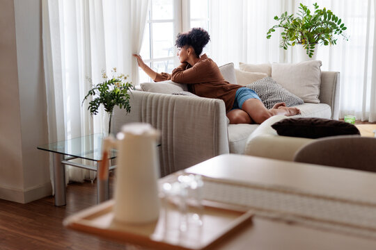 Woman gazes out window from sofa in bright room.