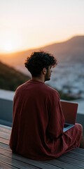 Fototapeta premium Young man sitting with laptop on terrace at sunset overlooking a coastal town for remote work background and travel lifestyle banner