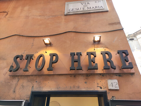 Rome, Italy - June 03, 2026: Stop Here sign mounted on a textured orange wall, illuminated by two lights, with a street name sign visible above in a historic urban setting