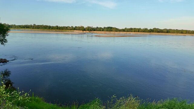 River flowing past green grassy bank toward tree lined opposite shore. Vistula