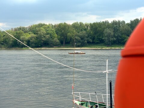 A scenic view of a wide river with a small motorized boat cruising in the distance. The foreground features details of a docked vessel with a rope, and a blurry orange object on the side, against a lu
