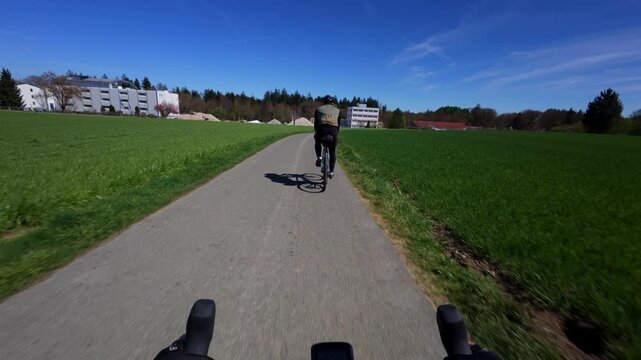 First person pov shot from drop handlebars tracking a road cyclist from behind on an empty asphalt countryside lane surrounded by vibrant green spring fields under a clear blue sky in Bavaria