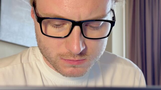 Close-Up of a Young Man Working on a Laptop, Wearing Glasses with the Screen Light Reflecting in His Lenses