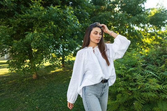 A young attractive woman with long brunette hair looking away thoughtfully in a sunny park. She wears a white linen boho blouse and denim jeans, posing gracefully by green bushes.