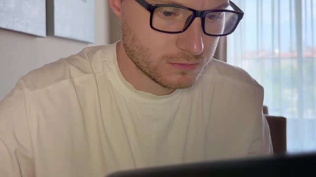 Close-Up of a Young Man Working on a Laptop, Wearing Glasses with the Screen Light Reflecting in His Lenses