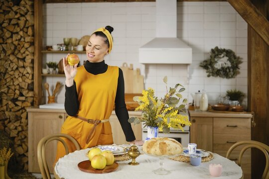 Cheerful woman standing by the kitchen table, holding a juicy apple in hand