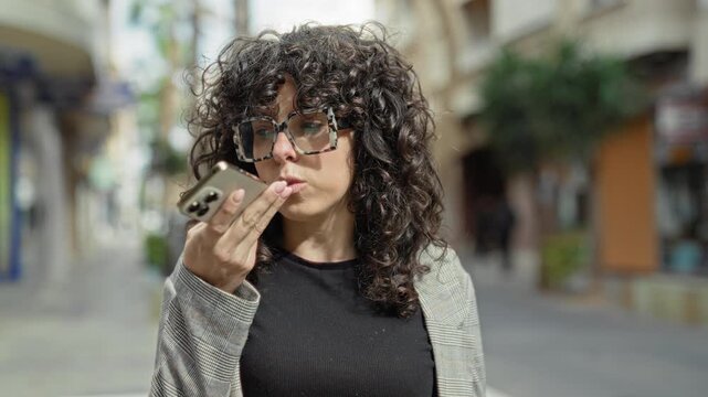 Woman holds smartphone to mouth speaking a voice message on a city street, wearing a checked blazer and oversized glasses; contemplation.