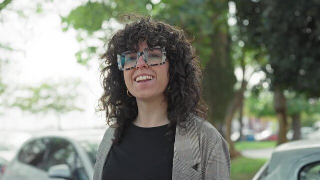 Young woman smiling and waving hand, wearing oversized tortoiseshell glasses and checked blazer on street; cheerful confidence.