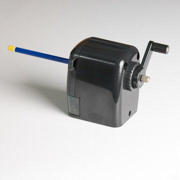 A classic black crank-handle pencil sharpener with a blue pencil inserted, ready for use. Positioned on a clean white background, highlighting its traditional design and functionality.