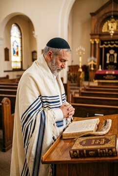Senior Jewish man in tallit and kippah praying with a shofar and prayer books in a traditional synagogue