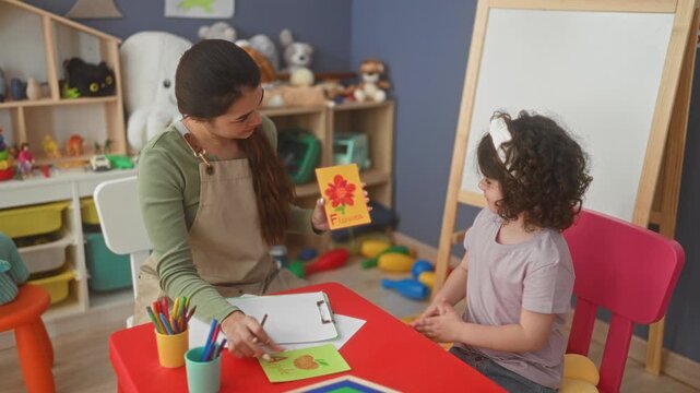 Woman teacher shows a flower flashcard and points with pencil to child's hand in preschool building; early learning joy.