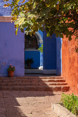 Fototapeta premium A stone pathway and steps lead past contrasting red and vibrant blue sillar stone walls under overhanging tree branches, looking toward a courtyard entrance inside the historic Santa Catalina Monaster