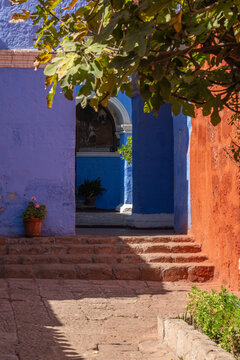 A stone pathway and steps lead past contrasting red and vibrant blue sillar stone walls under overhanging tree branches, looking toward a courtyard entrance inside the historic Santa Catalina Monaster