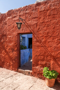 An open doorway in a vibrant red sillar stone wall leads into a contrasting blue courtyard decorated with potted plants at the historic Santa Catalina Monastery in Arequipa, Peru.