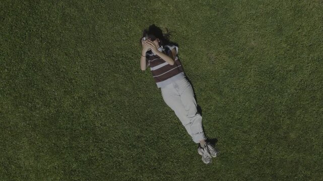 An aerial flyover of a young woman looking at her phone, laying on the grass in a park, St. George, Utah