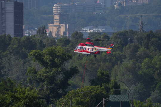 Firefighting helicopter of State Emergency Service hovering over green trees to refill water bucket from a lake in Kyiv Ukraine