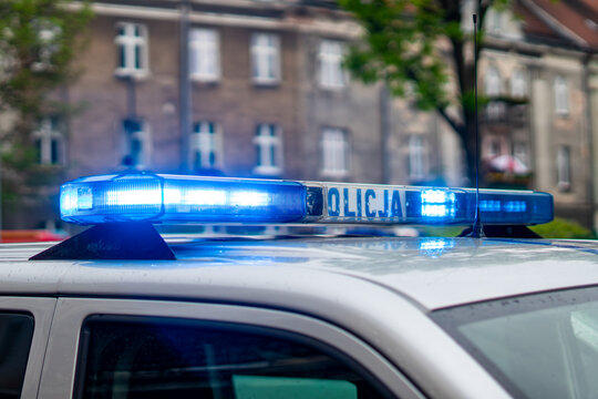 Blue police lightbar with white POLICJA lettering mounted on the roof of a silver patrol vehicle flashing bright LED emergency lights against a blurred urban building and green tree.