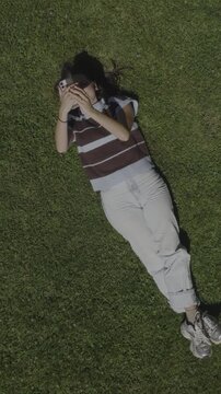 An aerial flyover vertical video of a young woman looking at her phone, laying on the grass in a park, St. George, Utah