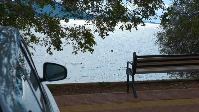 Parked car with the sea in the background. Serene coastal landscape featuring solitary bench overlooking calm blue waters, parked vehicle nearby, inviting peaceful moment of quiet reflection