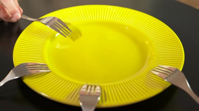 People placing forks around an empty plate. Several people simultaneously placing their forks around an empty plate on a black table, symbolizing concepts of waiting for food, collective hunger