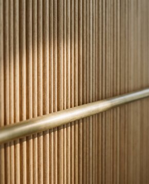 Close-up of fluted oak wall paneling with brass handrail in warm daylight, interior material detail