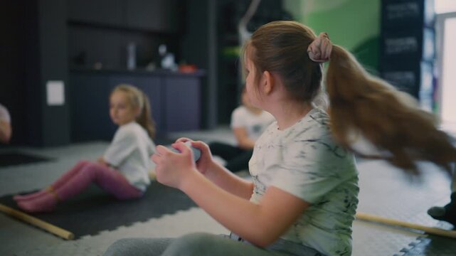 A focused young girl performs an exercise using a light blue prop during a children's fitness class. Several children engage in various physical activities in a gym setting.