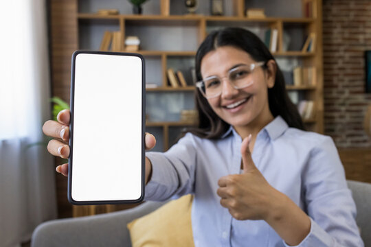 Young woman holding smartphone with blank white screen for copy space, giving a thumbs up gesture, smiling and looking at camera, representing good feedback, approval, and recommendation