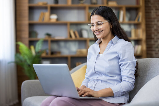 Young woman working remotely on her laptop computer from a cozy couch, wearing glasses and a blue shirt, smiling while engaging in online business tasks from home
