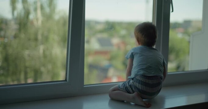 Little toddler boy sitting on windowsill and looking out the window