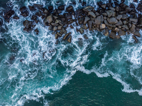 Aerial view of turquoise ocean waves crashing against dark jagged rocks with white sea foam along the coastline in Los Angeles, California, United States.