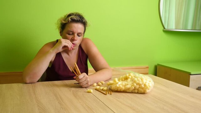 Thoughtful woman eating breadsticks and corn puffs while sitting at a table at home and snacking on unhealthy food