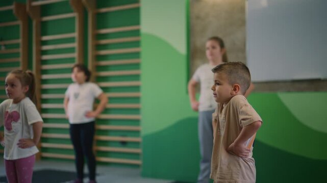 Young children perform fitness exercises and warm-ups in a gym. Kids participate in a group physical activity session.