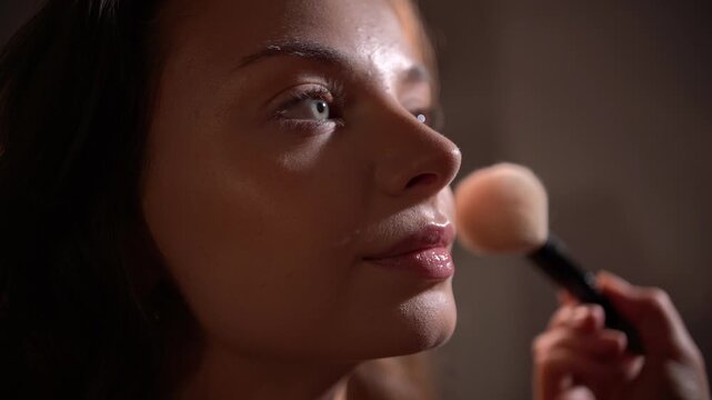 Professional makeup artist applying loose powder with large fluffy brush on model's face in close-up, model smiling and turning face from side to side, focus shift, warm backlight.

