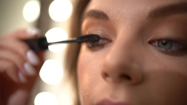 Extreme close-up of attractive young woman with blue eyes applying black cosmetic mascara to her upper eyelashes, carefully separating each lash with brush for perfect, glamorous look.
