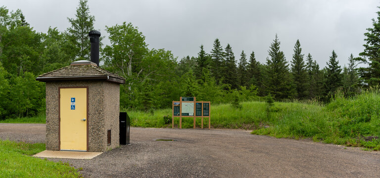Clean looking modern outhouse in a northern park setting next to a gravel parking lot with out of focus sign in the background
