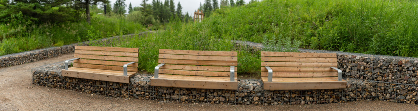 Panorama of 3 new wooden park benches in a natural park setting with a gabion wall.
