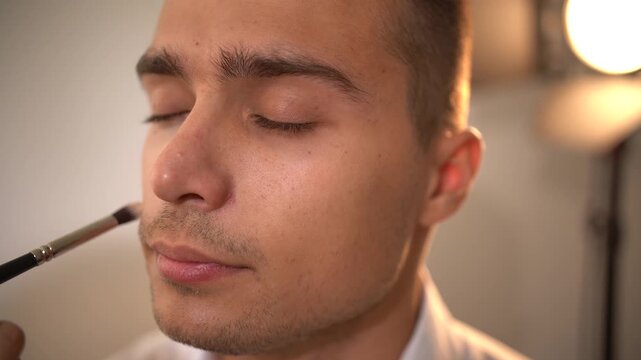 Professional makeup artist woman using small brush to apply concealer to handsome young man's left side of face in close-up profile view, model looking up then closing eyes.
