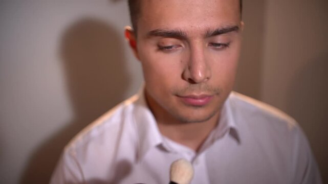 Professional makeup artist woman using large fluffy brush to apply powder to young man's face from above angle, model's eyes opening and closing, clean background.