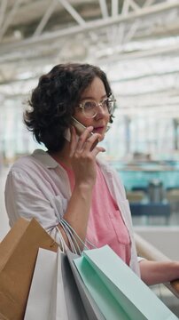 Active young woman talks on her phone while browsing summer clothing in store, looking through glass window display and holding paper shopping bag, a fashion lover engaged in a shopping conversation