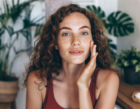 Woman with Freckles and Curly Hair Applying Skincare Near Green Plants