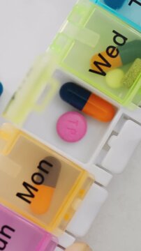 Close-up shows a man reaching into a pill organizer marked with the days of the week, extracting a medication tablet together with vitamins and capsules, part of a daily health management routine