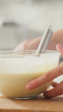 Close-up shows a woman's hands gripping a metal whisk, beating raw yellow eggs in a clear glass bowl illuminated by bright sunlight, part of the dough-mixing process as flour is added